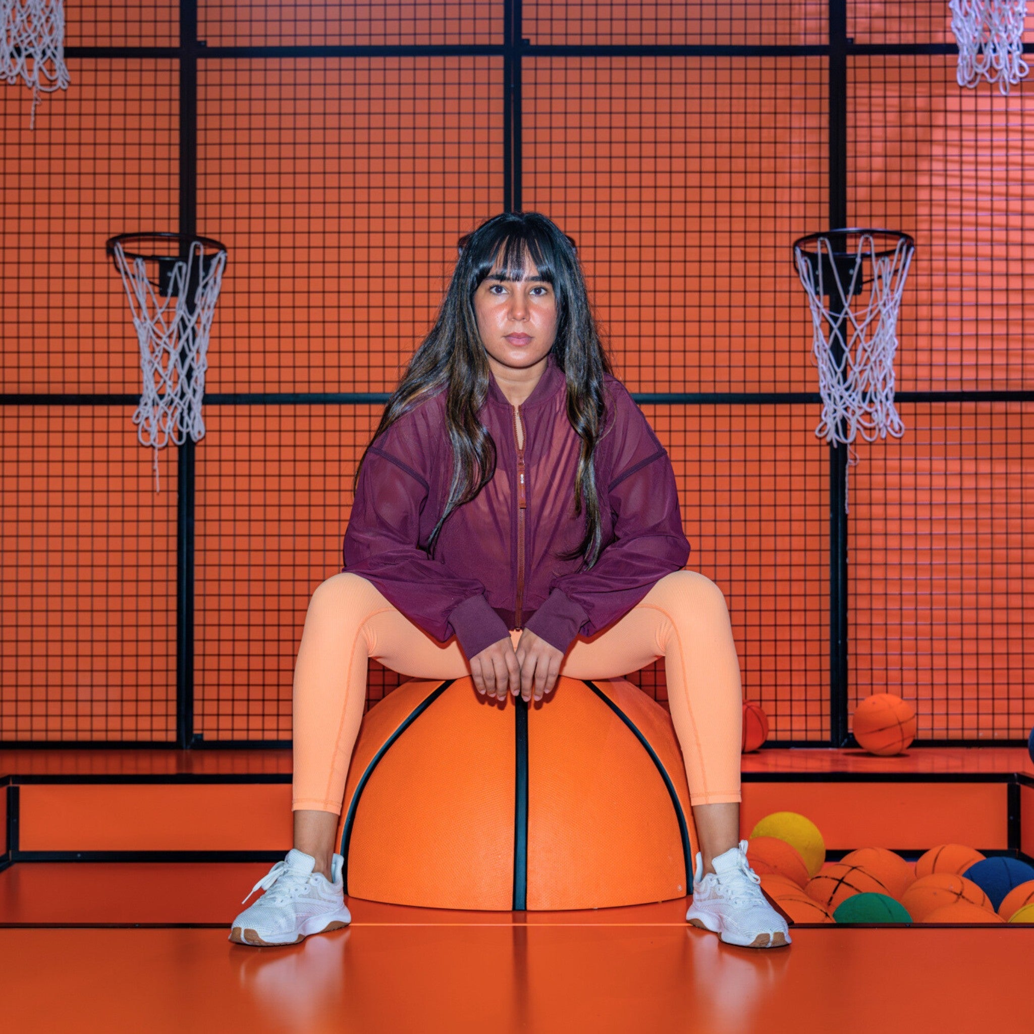A person wearing a maroon mesh jacket sitting on an orange basketball, with a basketball hoop and net in the background, and various colorful balls nearby.