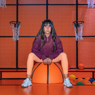 A person wearing a maroon mesh jacket sitting on an orange basketball, with a basketball hoop and net in the background, and various colorful balls nearby.