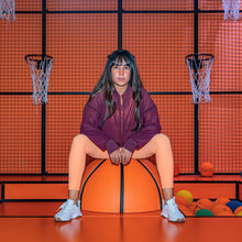 A person wearing a maroon mesh jacket sitting on an orange basketball, with a basketball hoop and net in the background, and various colorful balls nearby.