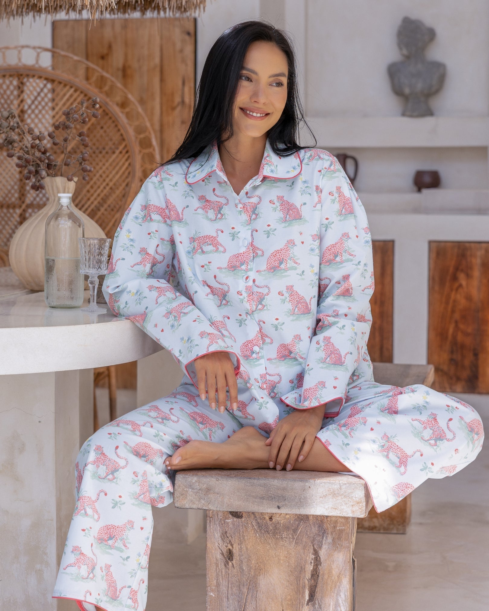 Layla Blue Long PJ Set – model seated on wooden stool smiling, wearing pale blue long-sleeve button-down pyjama shirt and trousers with pink LEOPARD and floral print and pink piping trim
